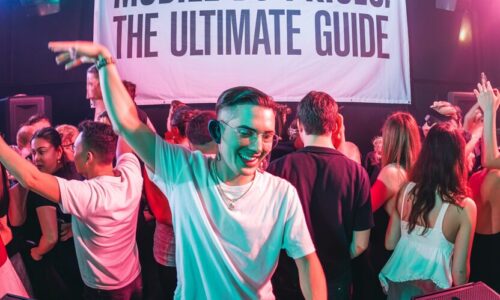 A smiling professional DJ with glasses performing at a vibrant event with a banner behind him reading 'MOBILE DJ PRICES: THE ULTIMATE GUIDE